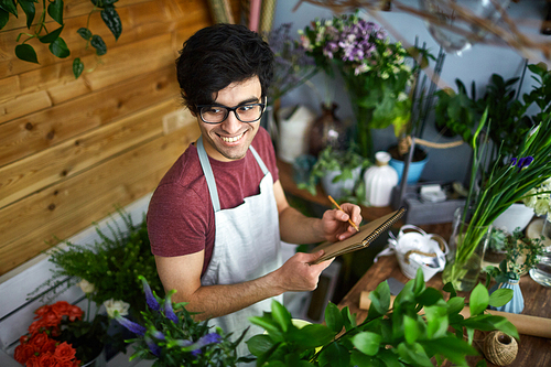 Young man in apron makingnotes in notepad in flower-shop