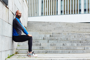 Sporty guy in headphones leaning against wall while exercising outdoors