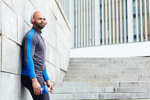 Guy in sportswear listening to music in headphones while leaning against wall