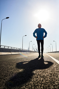 Outline of sportsman in akimbo pose standing in the middle of urban road or racetrack on sunny day with his shadow in front