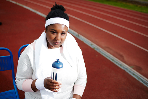 Portrait of plump African-American woman resting and drinking water after jogging on track