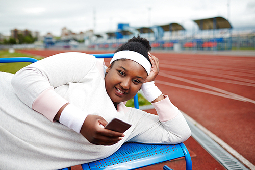Happy plus-size African-american girl in white sweatshirt, wristbands and headband lying on bench at stadium and texting in smartphone