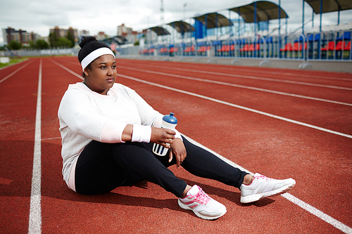Overweight African woman in sports clothing sitting on running track, recovering after jogging practice and thinking