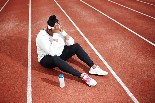 High angle view of frustrated overweight woman in sportswear sitting on running track with her head in hands, feeling exhausted