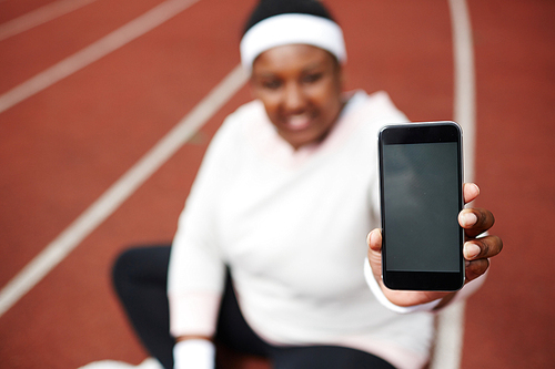 Defocused African overweight woman showing screen of her black smartphone while sitting on running track