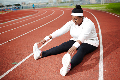 Plump sporty girl in white sweatshirt, cross-shoes and black leggins doing forward bends while sitting on racetrack with stretched legs