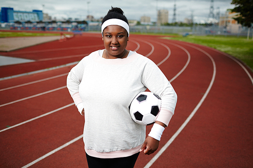 Portrait of positive plump woman posing with soccer ball at track and field stadium