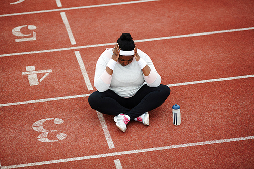 Exhausted overweight African woman sitting cross-legged near starting line on running track