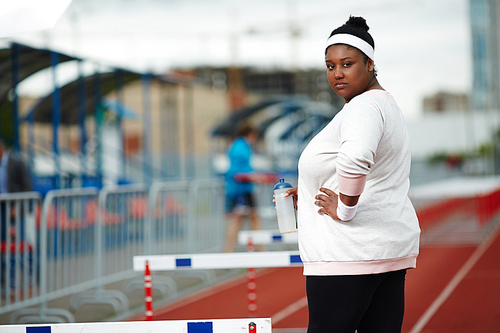 Fat female with bottle of water standing between hurdles on stadium