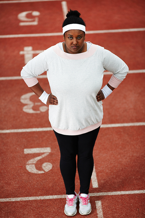 Portrait of overweight African-American woman in sportswear standing on running track with her hands on hips and 