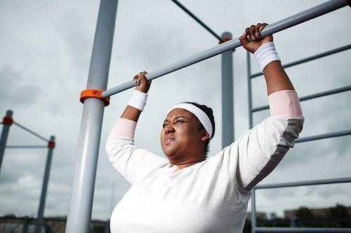 Persistant chubby girl trying to do difficult exercise while hanging on bar of outdoor sport facilities