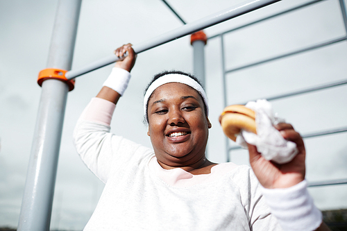 Happy young obese female with hamburger holding by bar of outdoor sport facilities and going to eat