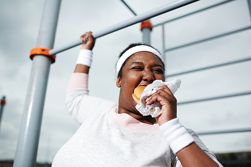 Very hungry young overweight woman eating sandwich while holding by bar of outdoor sport facilities during physical exercise