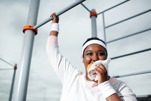 Plump African woman eating unhealthy burger while practicing pull-up exercise