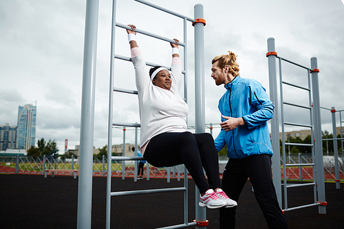 Overweight African woman practicing hanging double leg raise guided by personal trainer