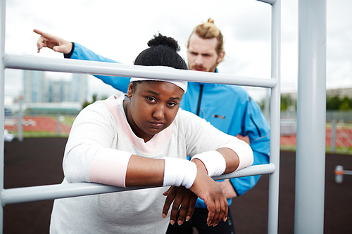 Portrait of tired chubby woman leaning on pull-up bar being motivated by personal trainer to keep exercising