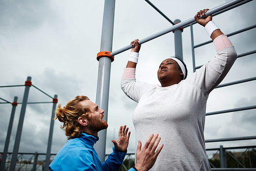 Determined oversized female hanging on facilities bars while trainer encouraging her