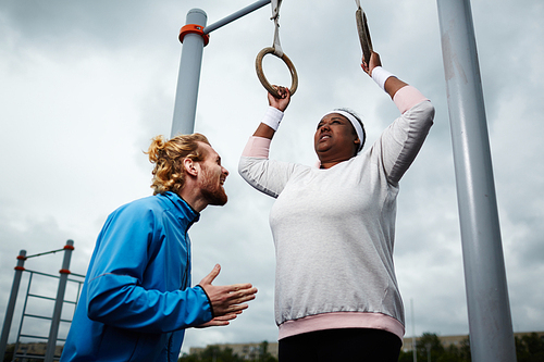 Experienced personal trainer motivating plus-size young woman to keep doing pull-up exercise on gymnastic rings