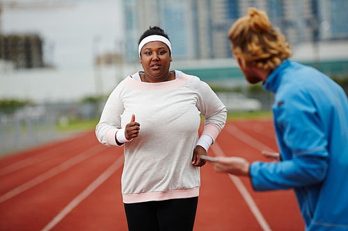 Determined plus-size woman running on track while personal coach measuring her time on smartphone