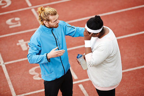 Personal trainer consoling tired overweight client after running practice at track and field stadium