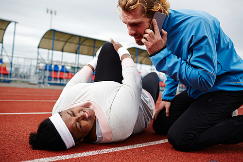 Plump woman with injured leg lying on running track, her trainer comforting her and  calling emergency service