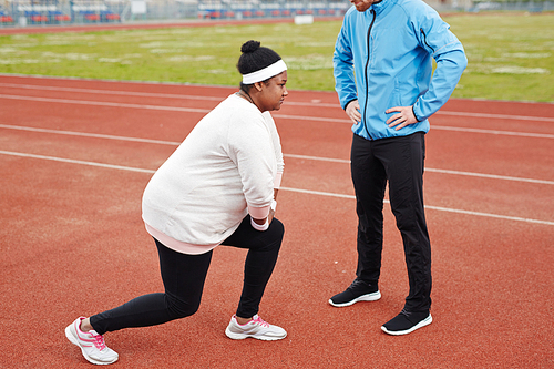 Young plump woman in activewear bending her knees during physical exercise with her trainer standing in front