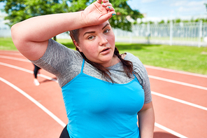 Chubby girl in sportive outfit sweating and touching her forehead during workout on stadium