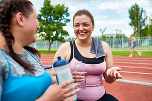 Friendly young women with bottles of water talking while having rest in the middle of workout on stadium