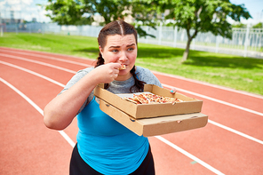 Hungry female in sportswear walking down racetrack and eating pizza from box