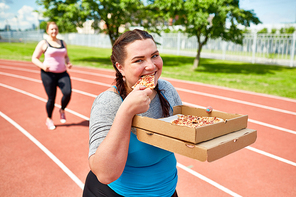 Young chubby woman with takeout pizza eating it on stadium after training