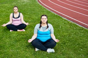 Two cross-legged chubby females practicing yoga on stadium