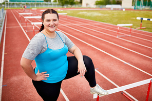 Successful overweight woman keeping one leg on hurdle while standing on racetrack