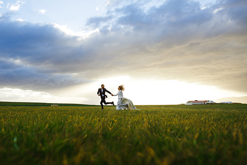 Bride and groom running down green pasture in rural environment