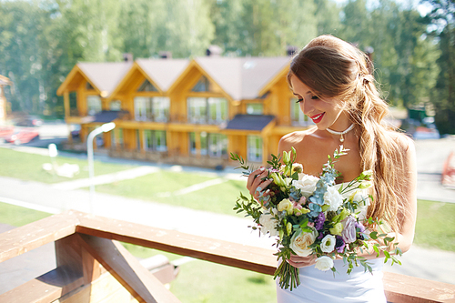 Happy young bride looking at floral bouquet on sunny day