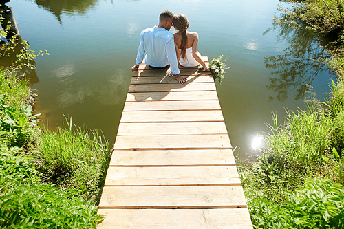 Backs of bride and groom sitting on pontoon by lake