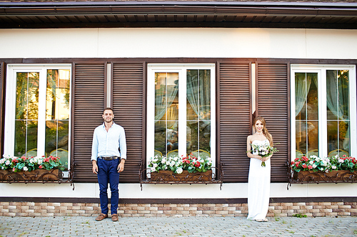 Smiling bride and groom standing by rural edifice with small flower-beds on windows