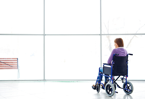 Rear view of handicapped young woman sitting in wheelchair, looking at window and daydreaming
