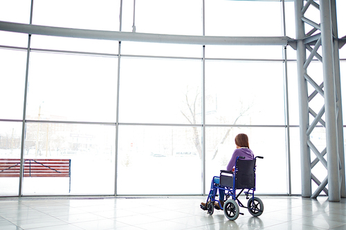 Rear view of disabled woman sitting in wheelchair in shopping mall and looking at big window