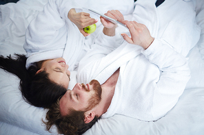 Modern couple in bathrobes using touchpad in bed