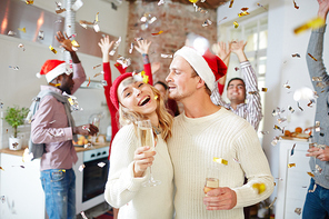 Laughing girl and her boyfriend in sweaters and caps enjoying xmas party with friends