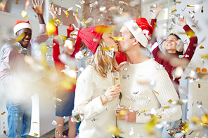 Young kissing couple with flutes of champagne standing in confetti fall at party