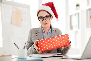 Young employee in Santa cap and formalwear holding big wrapped box while sitting by her workplace