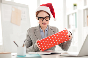 Happy young businesswoman looking at packed Christmas gift in her hands