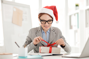 Smiling young impatient employee untying red ribbon on top of package with xmas gift