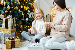 Little girl with xmas toy sitting by decorated firtree while her grandmother plating her hair