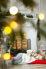 Adorable girl sleeping on the floor on red pillow by fireplace and decorated Christmas tree