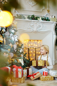 Adorable child with present looking at decorated firtree while sitting by fireplace at home on xmas night