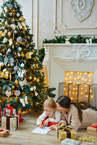 Grandmother and granddaughter lying on the floor by fireplace and decorated firtree and drawing xmas picture