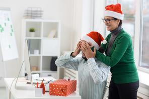 Laughing woman covering her co-worker eyes by hands while making Christmas surprise