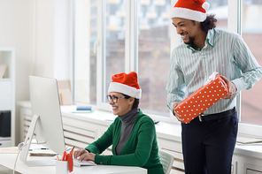 Happy man with giftbox and his co-worker looking at monitor in office on xmas day